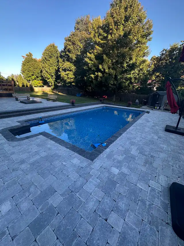 Finished pool with a grey interlock patio under an evening sky and evergreens