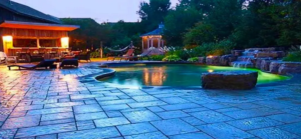 Freeform pool at dusk with a rock waterfall, a bar cabana and lit interlock