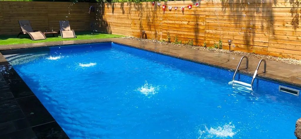 Rectangular pool with fountain jets arcing in, a wood fence and loungers